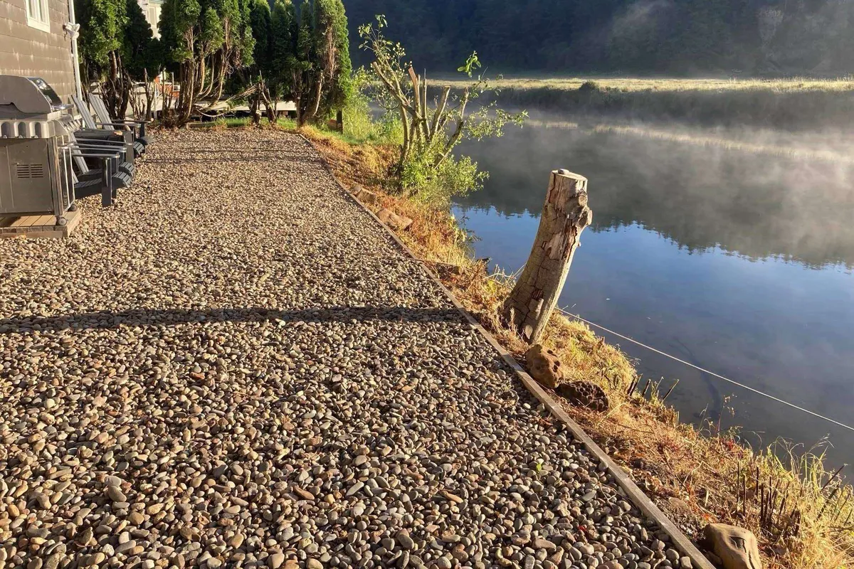A peaceful riverside scene featuring a gravel pathway, trees, a seating area, and the calm water surface. The natural setting is ideal for dog walks, nature exploration, and unwinding close to the water.