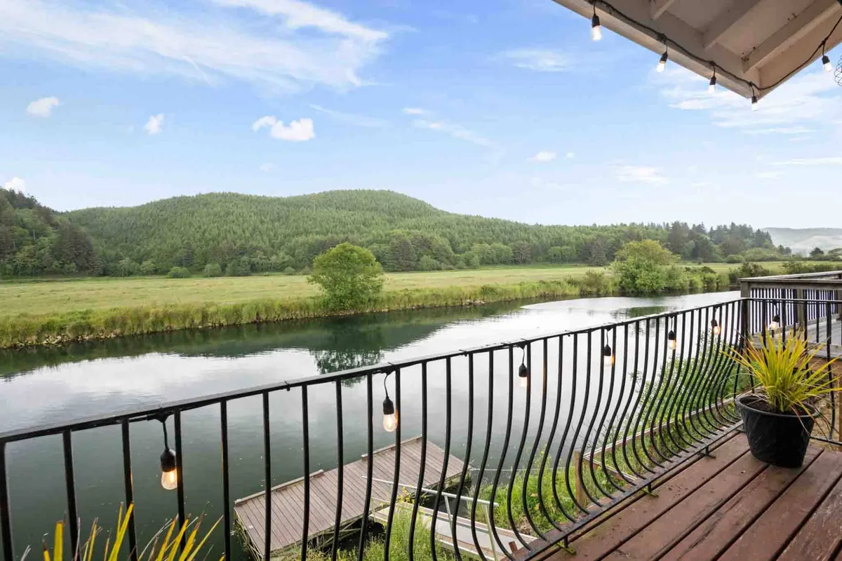 A serene exterior view overlooking the Nestucca River, with lush green hills in the background, a balcony railing in the foreground, and warm hanging lights creating an inviting atmosphere. The area is set up for relaxing, fishing, or enjoying a meal while watching for wildlife along the riverbanks.