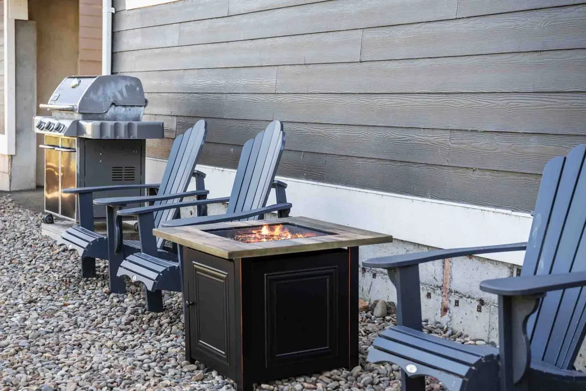 An inviting outdoor gathering space with a fire pit surrounded by Adirondack chairs and a grill nearby, set in a natural environment. The scene is ready for social gatherings, barbecues, and sharing stories as the sun goes down.