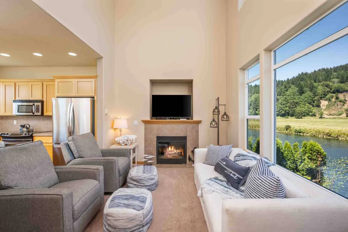 A sunlit living area with large windows framing a picturesque river view. Plush chairs and a cozy sofa are arranged around a gas fireplace, creating an inviting spot for relaxation. The abundant natural light and scenic outlook emphasize the connection to nature and wildlife just outside.