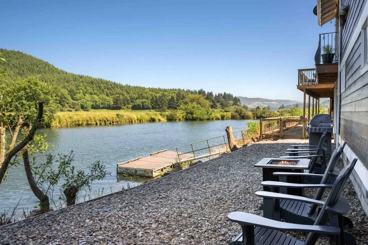 This image shows a spacious deck overlooking a serene river, with chairs arranged for relaxation. The landscape features lush greenery and gentle hills, while the water reflects the sky, creating a peaceful ambiance ideal for outdoor lounging or observing nature.