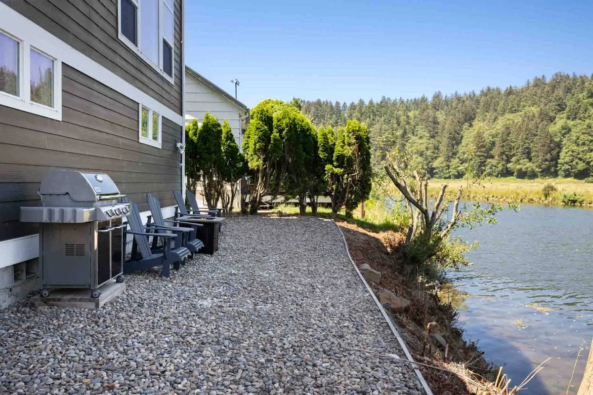 A riverbank scene with Adirondack chairs and a grill, set under the trees with the river flowing nearby. The atmosphere is casual and welcoming, suggesting a great spot for outdoor dining, play, and relaxation with pets.