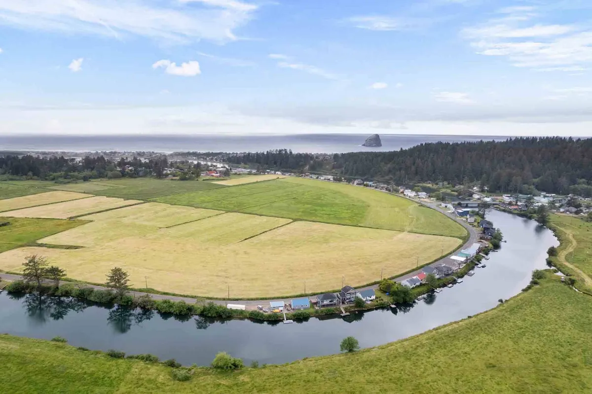 A tranquil, wide-angle view of the river with surrounding fields, a glimpse of the distant ocean, and several homes dotted along the riverbanks. The soft golden light suggests the time is around sunset, enhancing the romantic atmosphere. The peaceful waters and surrounding nature establish a serene backdrop ideal for a romantic toast or quiet evening.