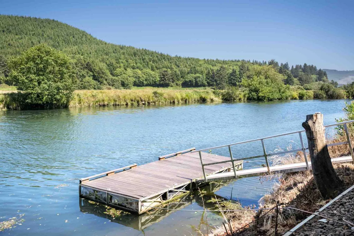 A wooden dock extends over calm water, surrounded by green foliage and backed by distant mountains. The setting feels peaceful and secluded, ideal for fishing, kayaking, or simply soaking in the natural beauty.