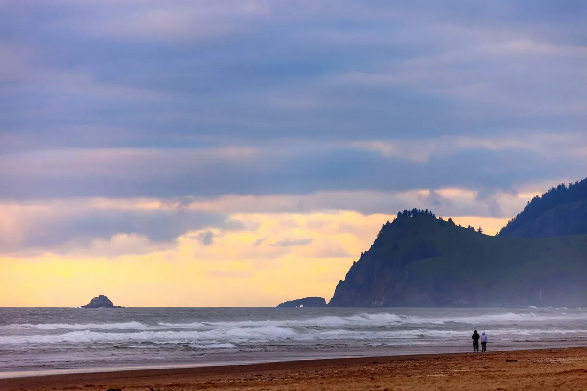A wide sandy beach with gentle waves, a couple walking in the distance, distant cliffs, and striking rock formations. The open beach offers plenty of space for dogs to run and families to enjoy nature.