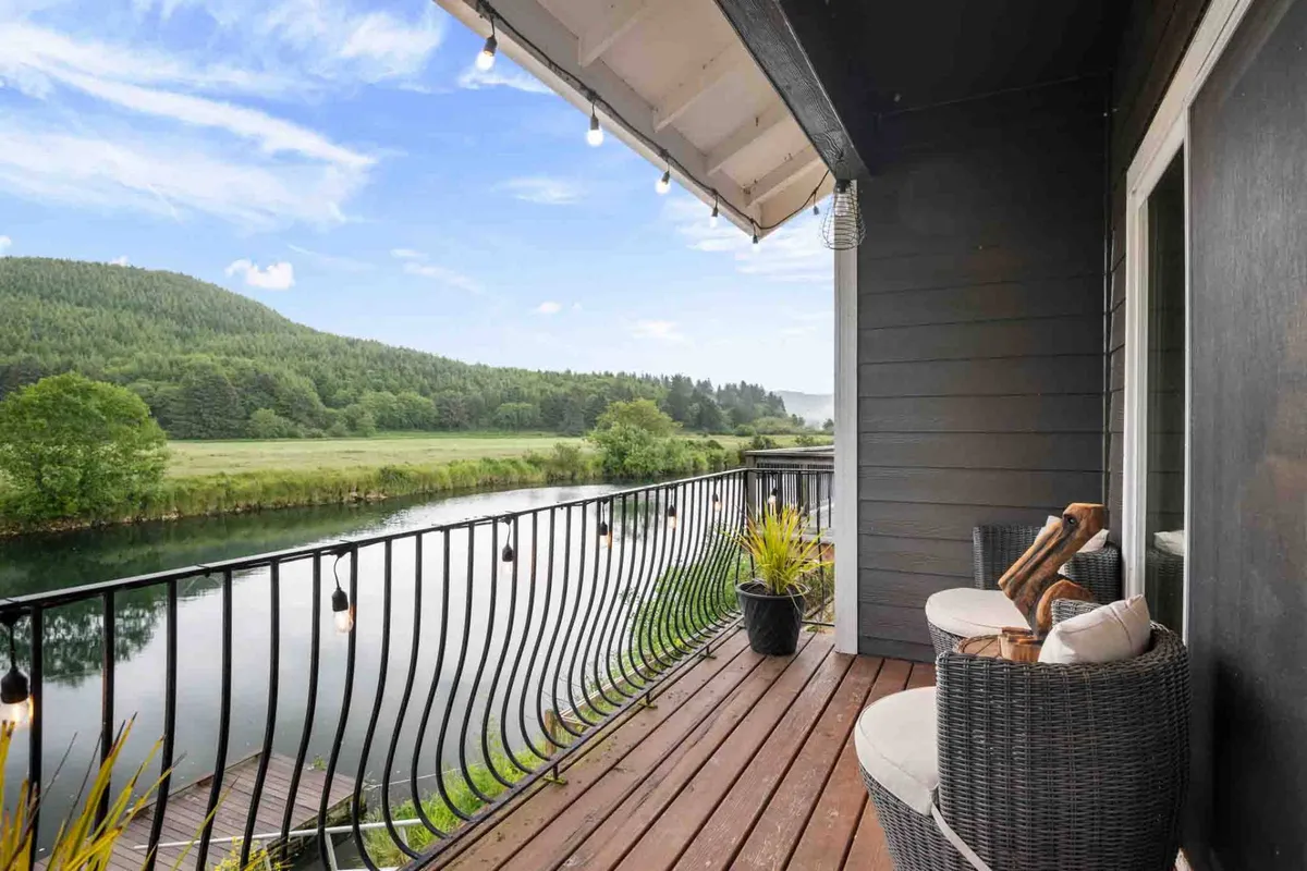 An exterior view from the deck or balcony with comfortable seating, potted plants, and sweeping river views. The setup invites al fresco dining, morning pastries, and wildlife watching just outside the main living areas.