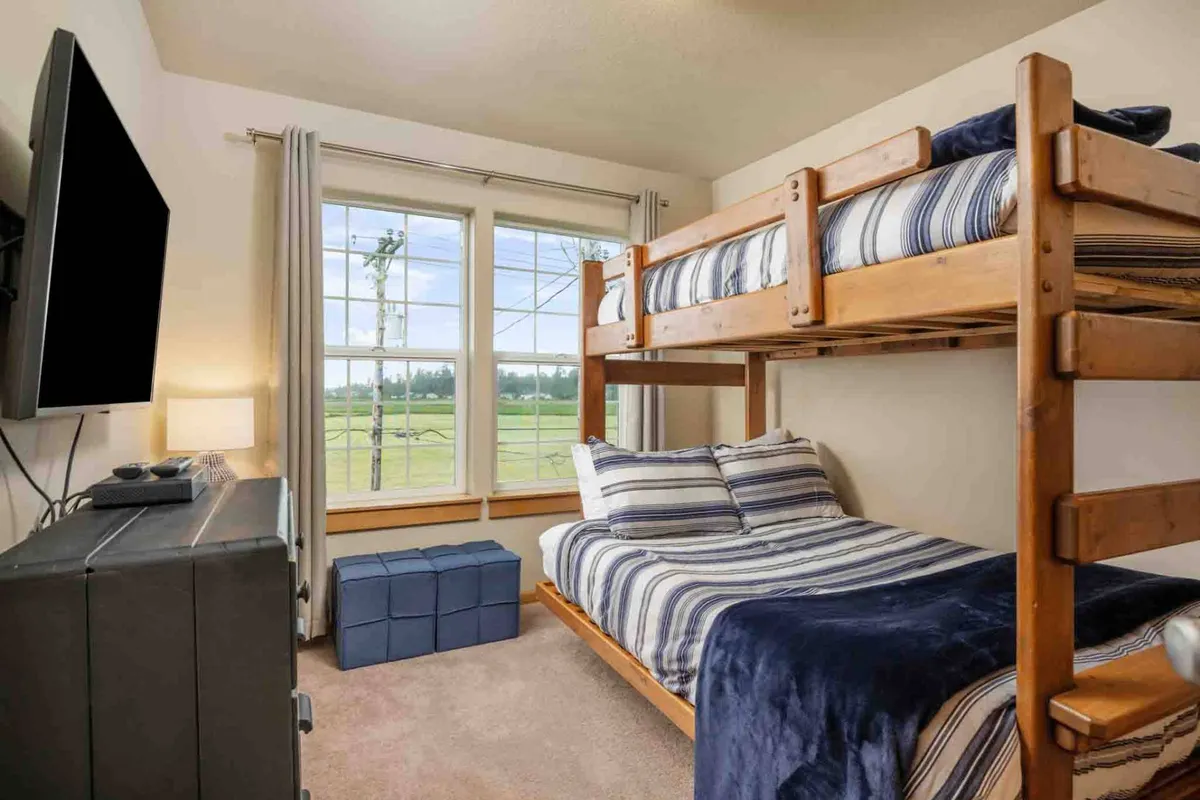 A playful bunk room featuring a bunk bed (double/twin), large windows that let in abundant natural light and offer field or ocean views, a television, and a blue storage ottoman for games or books. Warm, cozy, and ideal for children or teens.