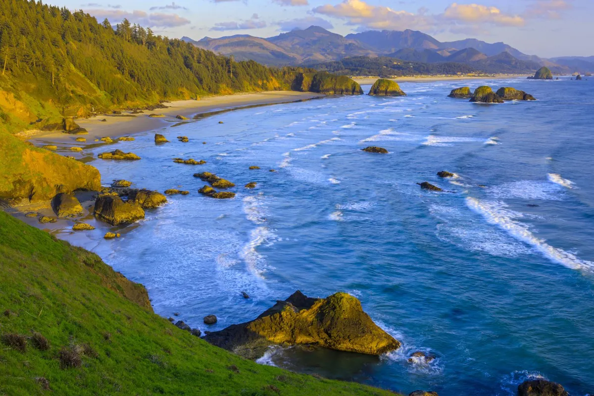 A sweeping view of the beach shoreline with ocean waves crashing against iconic rock formations, distant mountains in the background, and wide open sky above. The sand appears inviting for beachcombing and play, and the overall landscape embodies the wild beauty of the Oregon coast.
