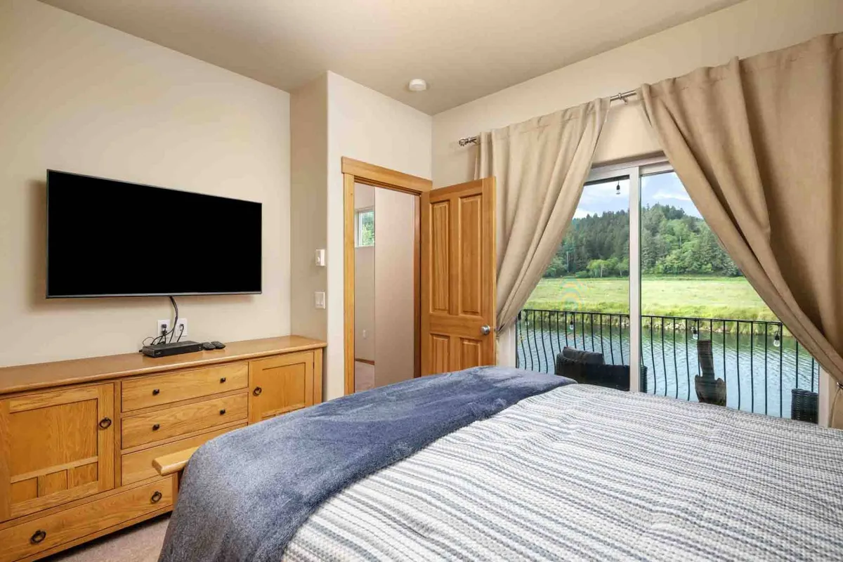 A cozy bedroom with a bed draped in a striped blue blanket, complemented by a large television and a wooden dresser. Curtains frame a window that reveals a scenic view, providing both comfort and connection to the outdoors.